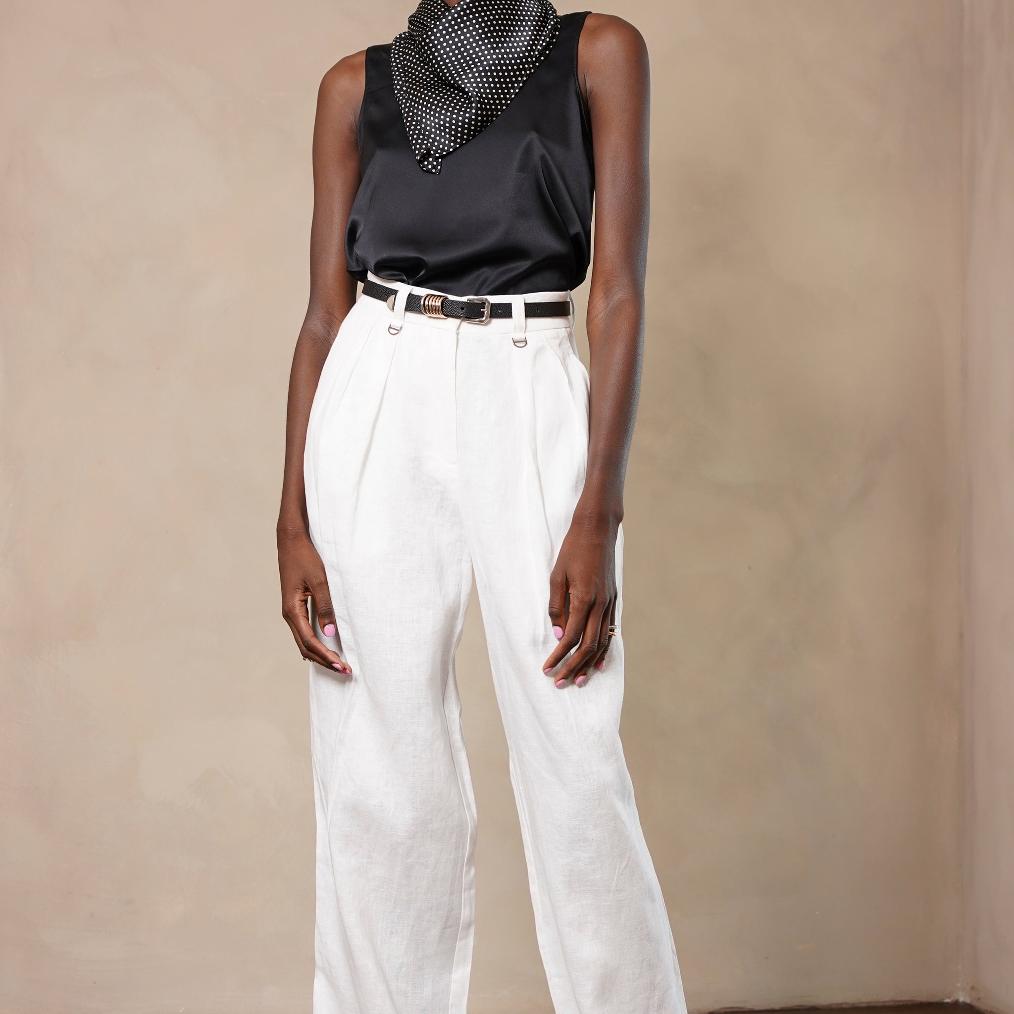 Model wearing a black sleeveless top and white pants against a beige wall.